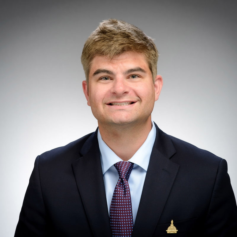 Headshot of Thomas Waldschmidt, a man with short light brown hair, wearing a navy suit, light blue shirt, and maroon patterned tie, smiling against a gray background. He wears a small gold University of Notre Dame lapel pin.